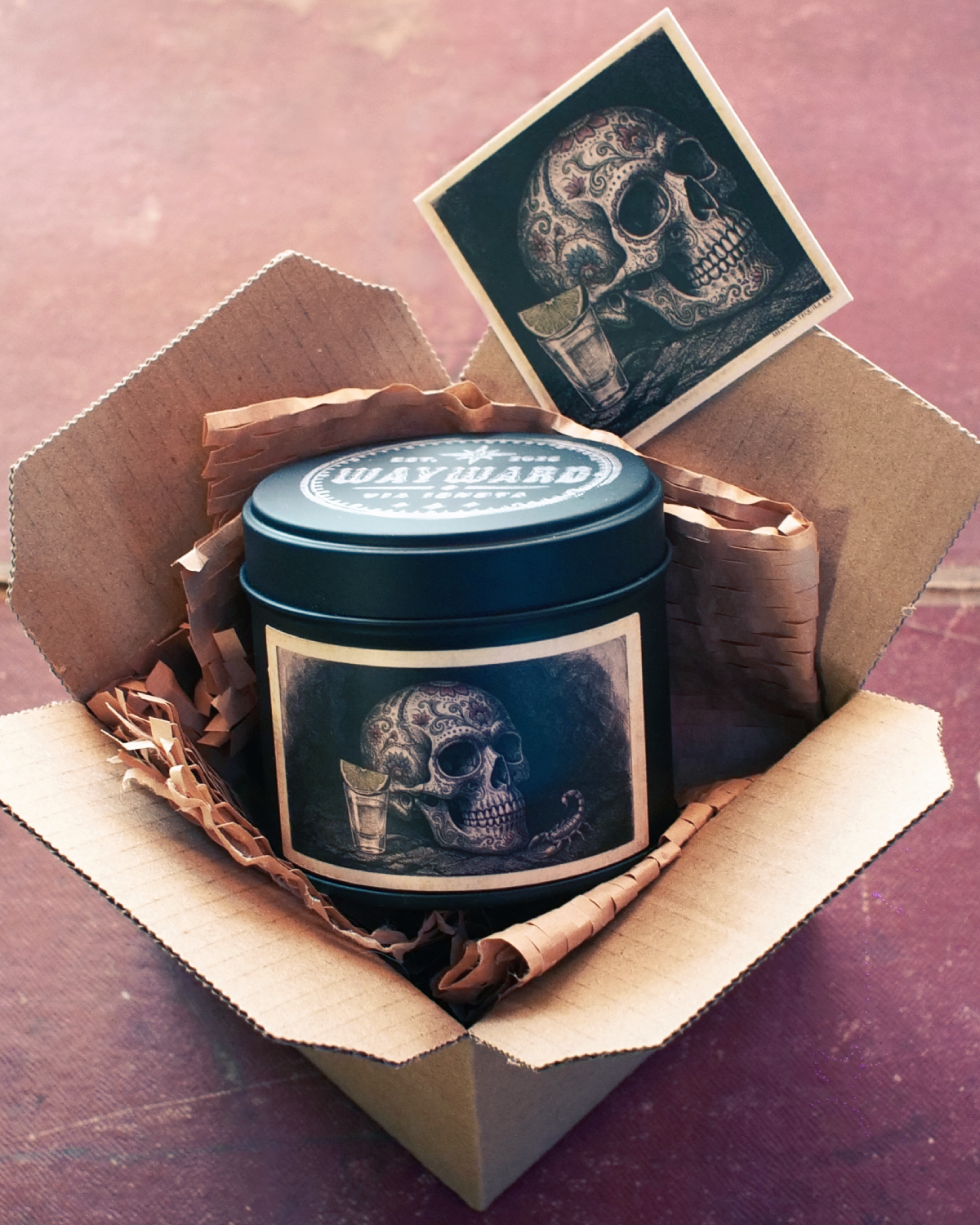 A dark metal tin labeled 'wayward' with a skull graphic, presented inside an open cardboard box with tissue paper and a matching skull card, on a reddish-brown surface.