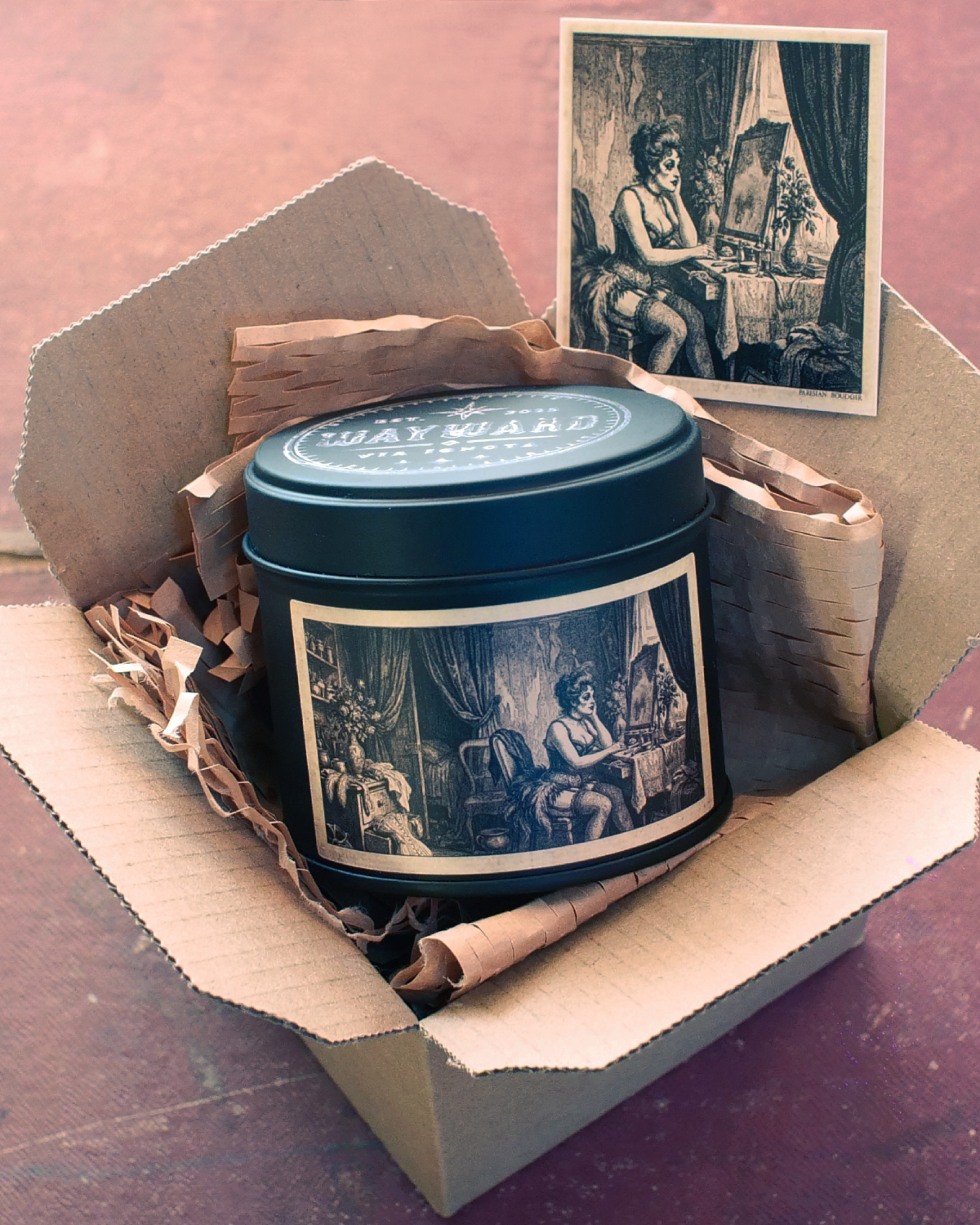 A dark teal metal tin candle with vintage-style black and white illustrations, presented inside an open cardboard gift box with shredded paper and a matching vintage print card on a reddish-brown surface.