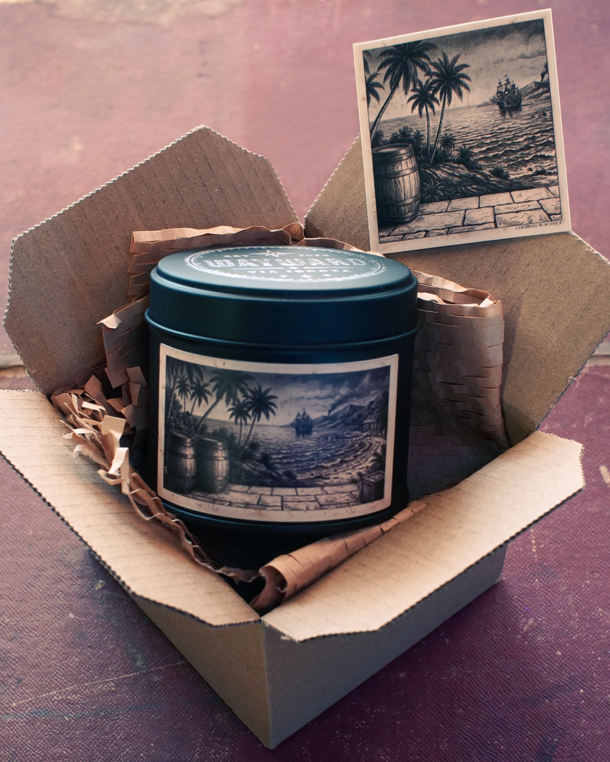 A dark teal metal tin with a vintage-style tropical beach illustration sits inside an open cardboard gift box with brown paper filler. A matching printed card is propped behind the tin on a textured mauve-pink surface.