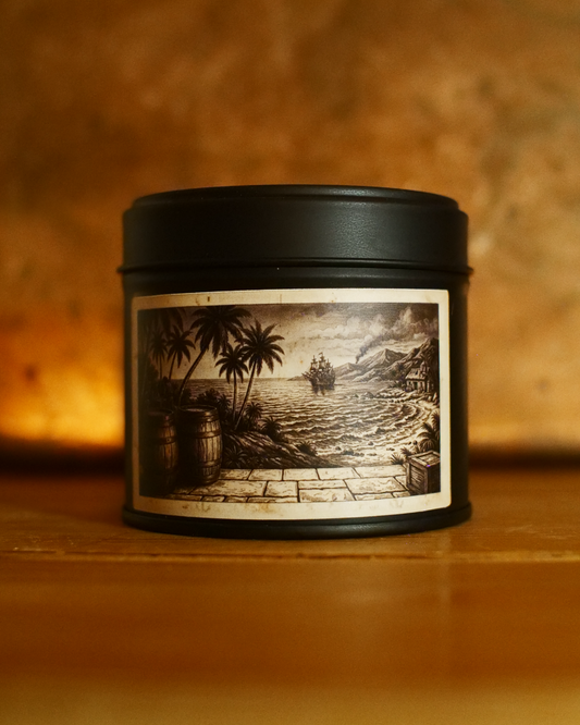 Caribbean Rum Shack candle in a matte black tin, lid on, with an etched label of rum barrels beneath palms, a stone quay and a tall ship on the horizon — Wayward