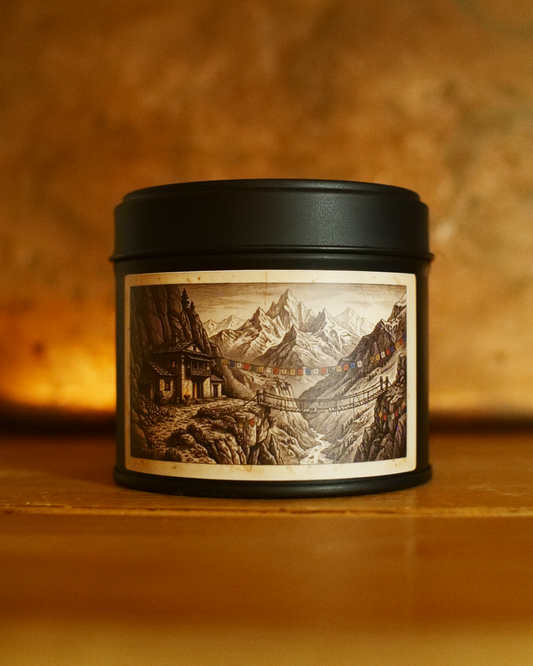 Himalayan monestary candle in a matte black tin, lid on, with an etched label of a mountain lodge, prayer flags and a rope bridge over a gorge — Wayward