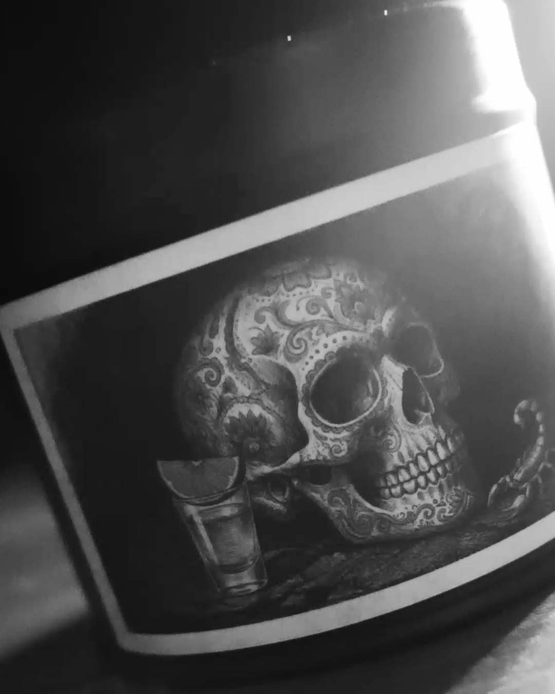 Black-and-white advert still: close, tilted view of the Mexican Tequila Bar tin, etched sugar-skull label filling the frame