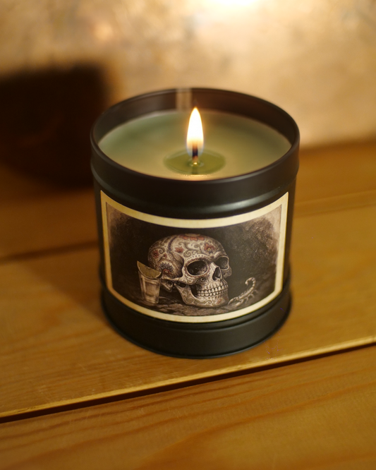 Mexican Tequila Bar Candle by Wayward. A black metal candle jar with a lit flame, featuring a vintage-style label with a skull illustration, set on a wooden surface against a warm, blurred background.