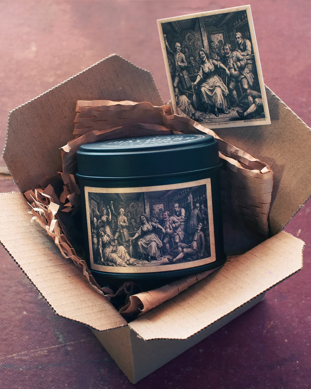 A dark teal metal tin with a vintage black-and-white illustration label, nestled inside an open cardboard box with brown paper filler. A matching small print card with a black-and-white classical scene lies above the tin on a reddish-brown surface.