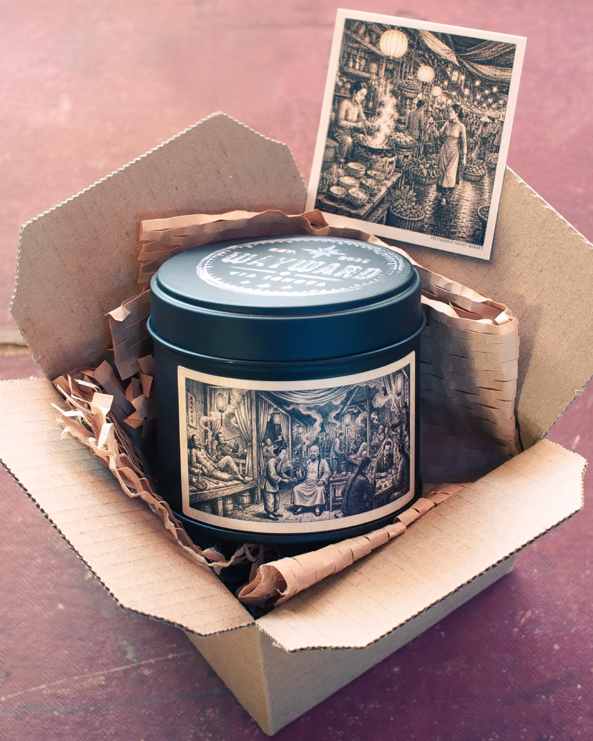 A dark teal metal tin with a vintage-style black and white illustration label, nestled inside an open cardboard box with crumpled paper filler and a matching illustrated card propped behind it.